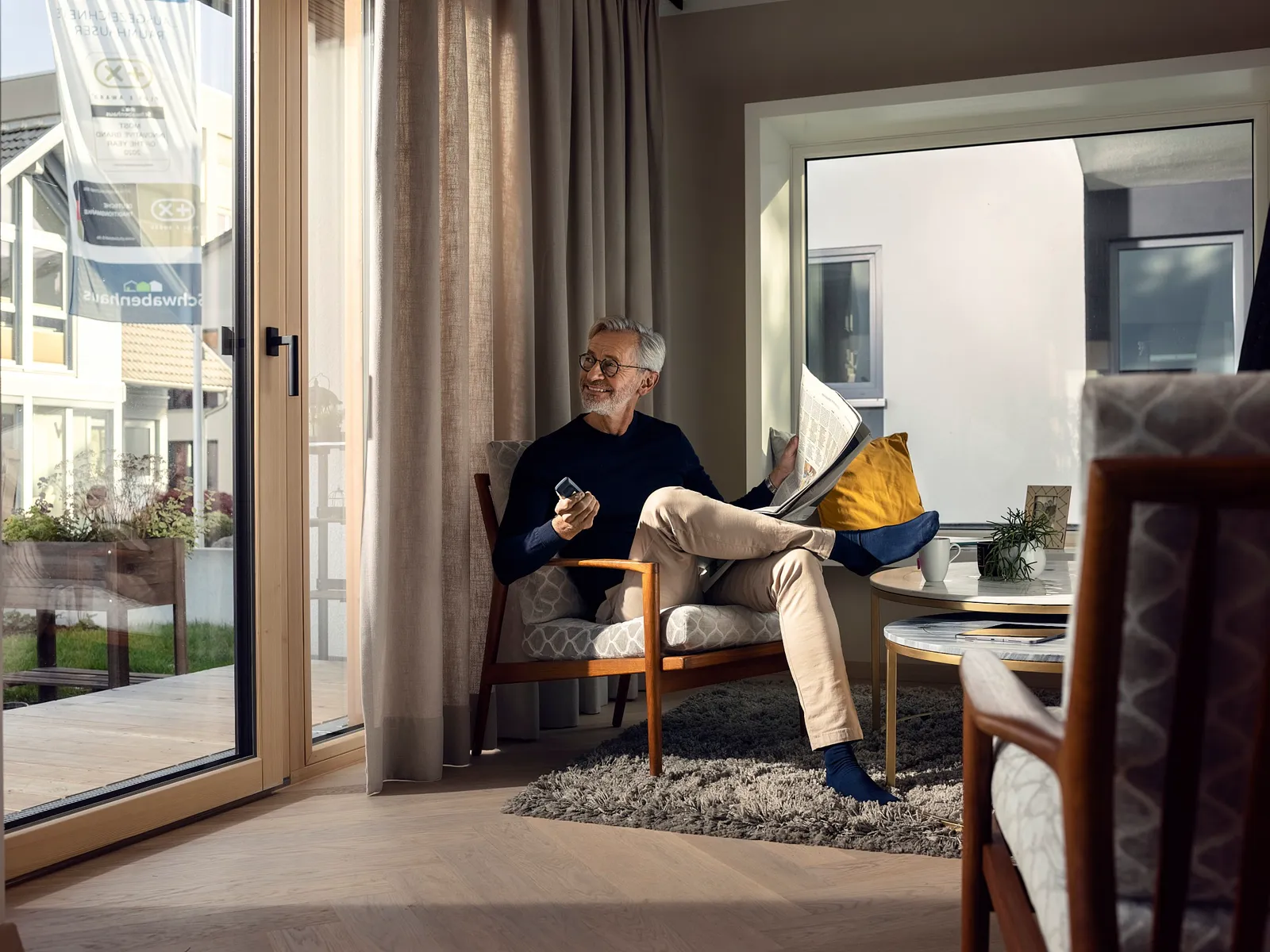 Mann liest Zeitung im Sessel am bodentiefen Fenster, Herbstlicht fällt herein